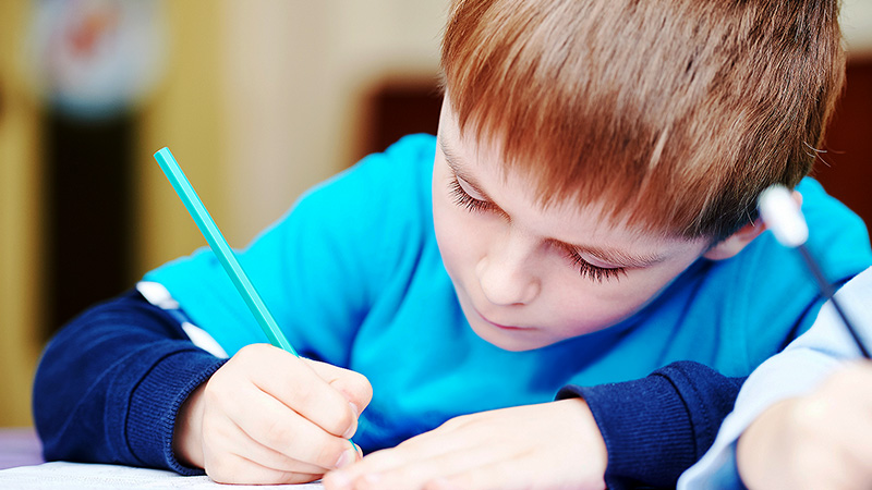 Young boy wearing blue focusing on writing on a piece of paper. 